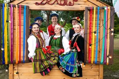 Grupa osób w strojach ludowych pozuje w drewnianej, kolorowo przystrojonej budce na zewnątrz.