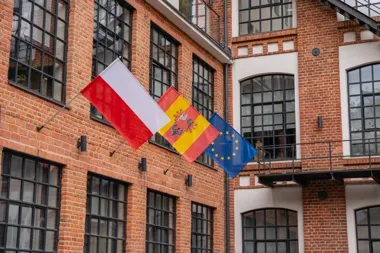Trzy flagi – polska, hiszpańska i unijna – wiszą na elewacji z czerwonej cegły przed budynkiem z dużymi oknami.