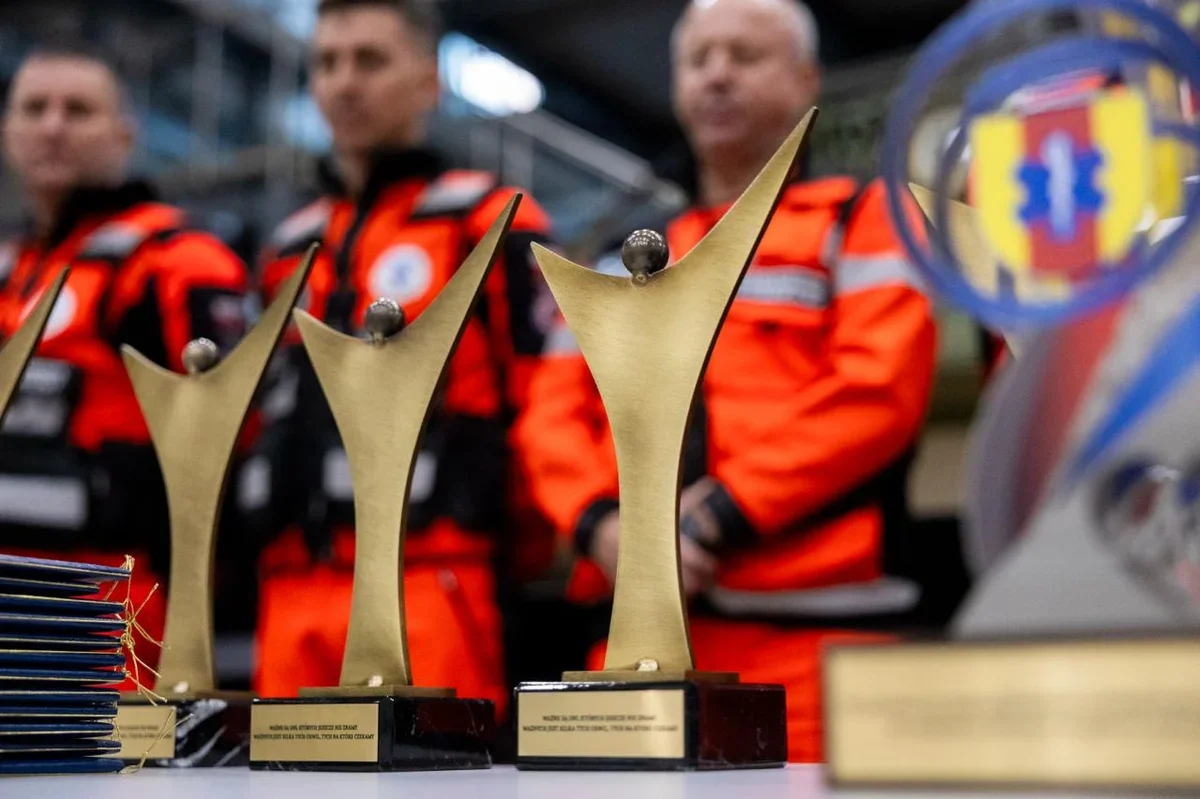 Trzy złote statuetki i dyplomy leżą na stole, a w tle stoją trzej mężczyźni w pomarańczowych mundurach ratowniczych.